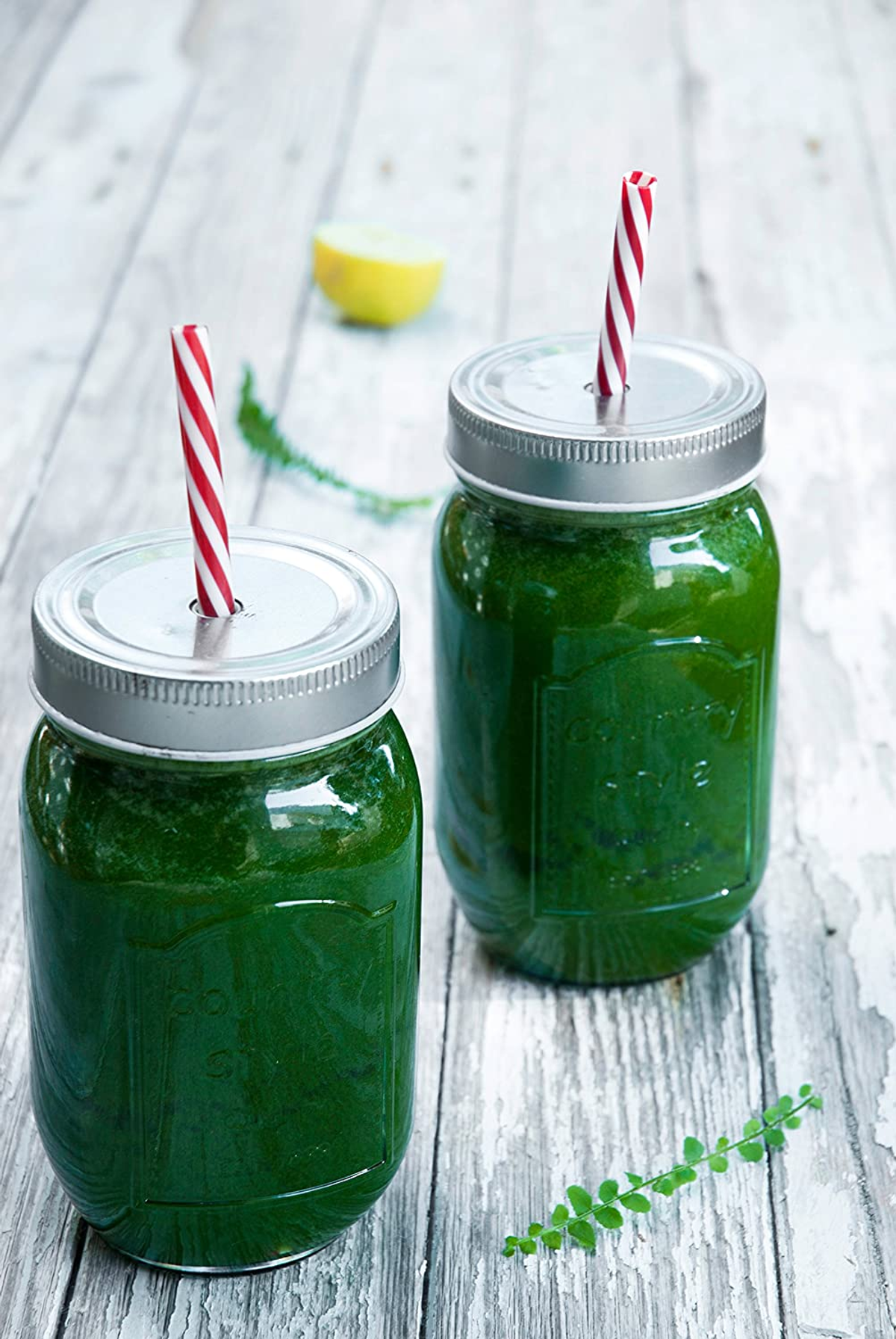 Dos batidos verdes en frascos con pajitas, rodaja de limón, hoja sobre madera blanca.