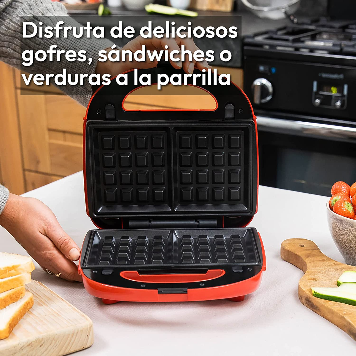 Waffeliera rossa aperta su un tavolo. C'è un piatto di pane e una ciotola di fragole.