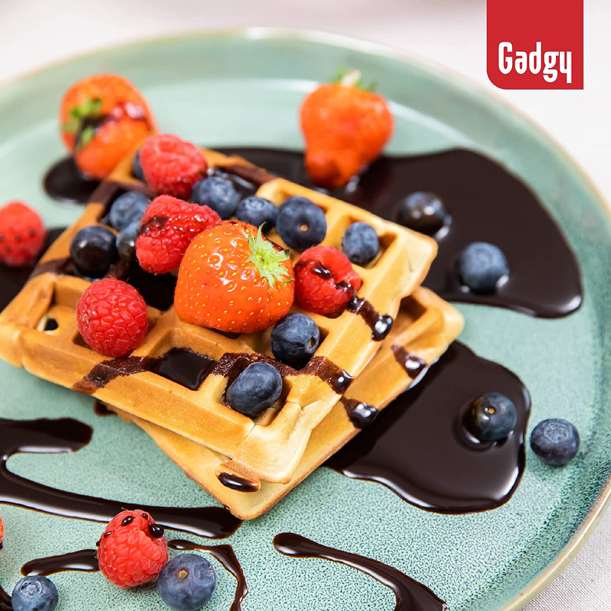 Waffle con fragole, mirtilli e sciroppo di cioccolato su un piatto verde. Logo Gadgy.