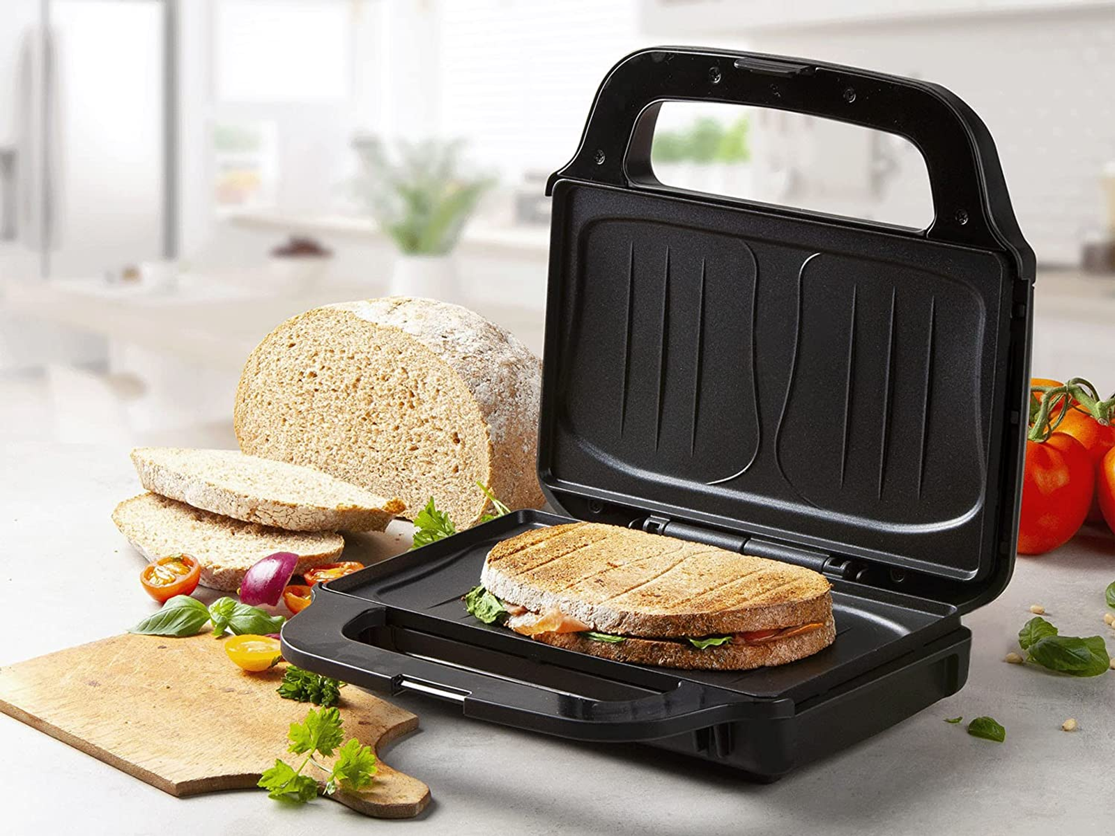 Kanapka w czarnej opiekaczu do kanapek. Chleb, składniki i drewniana deska do krojenia są w pobliżu.