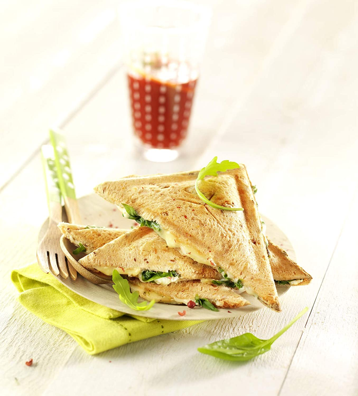 Sándwiches en un plato con hojas de rúcula. Tenedor de madera y servilleta verde. Bebida roja detrás.