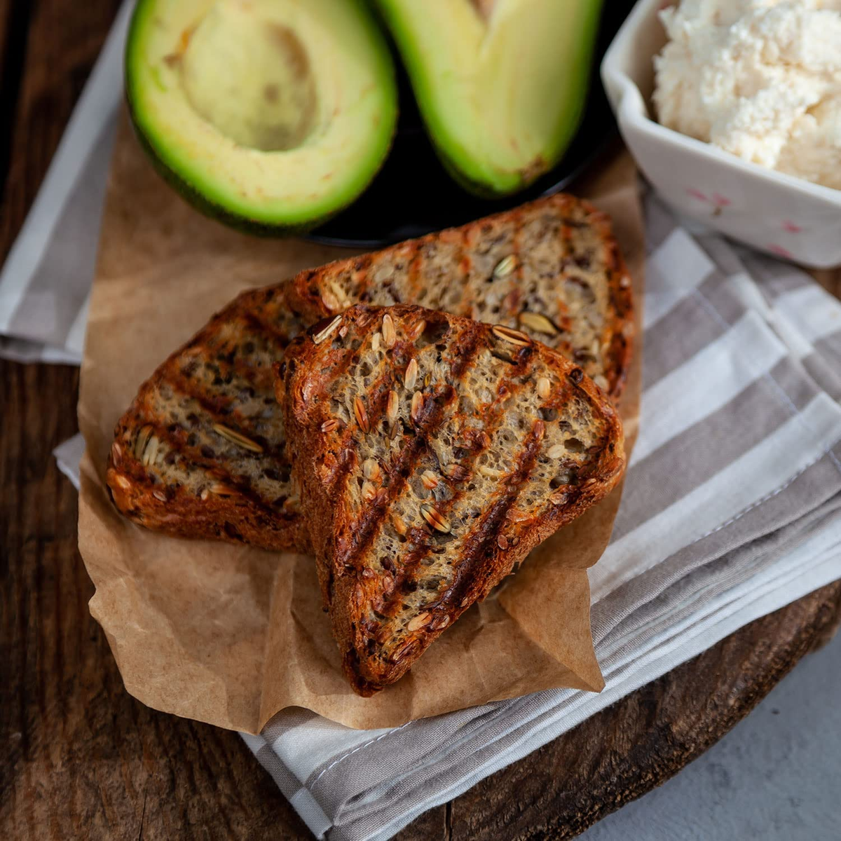 Rebanadas de pan a la parrilla con semillas, mitades de aguacate y un cuenco de queso blanco sobre una superficie de madera.