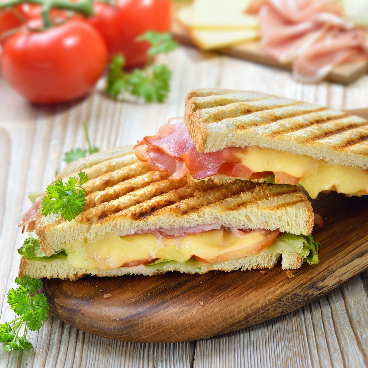 Dos sándwiches a la plancha con jamón, queso, tomate y lechuga en una tabla de madera, perejil al lado.