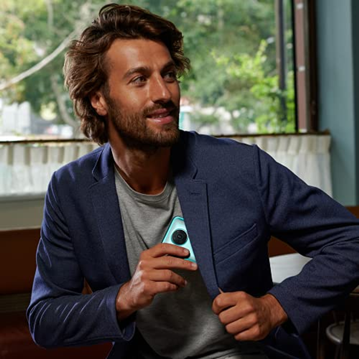 Een man in een blauwe jas houdt een blauwgroene telefoon in zijn zak, in een lichte kamer.