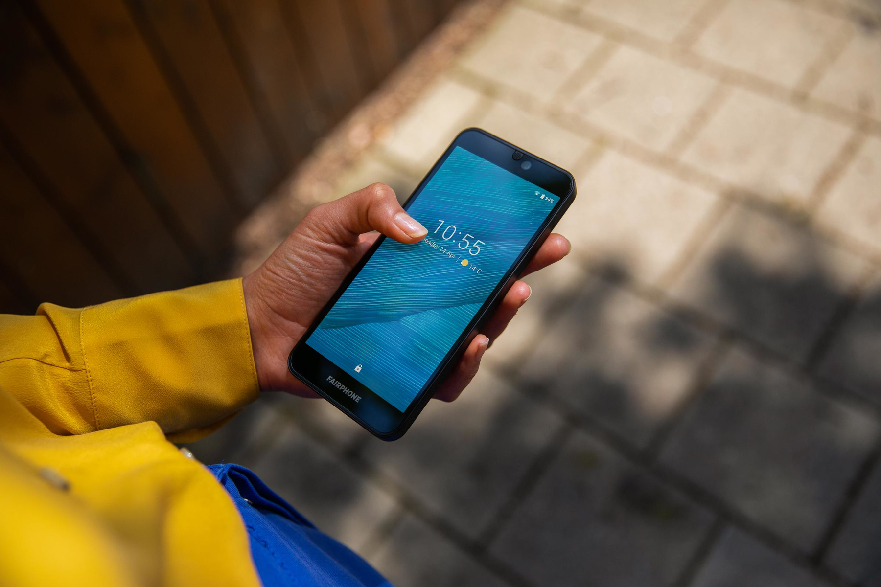 Hand hält Fairphone. Blauer Bildschirm zeigt Zeit und Wetter. Außenaufnahme.