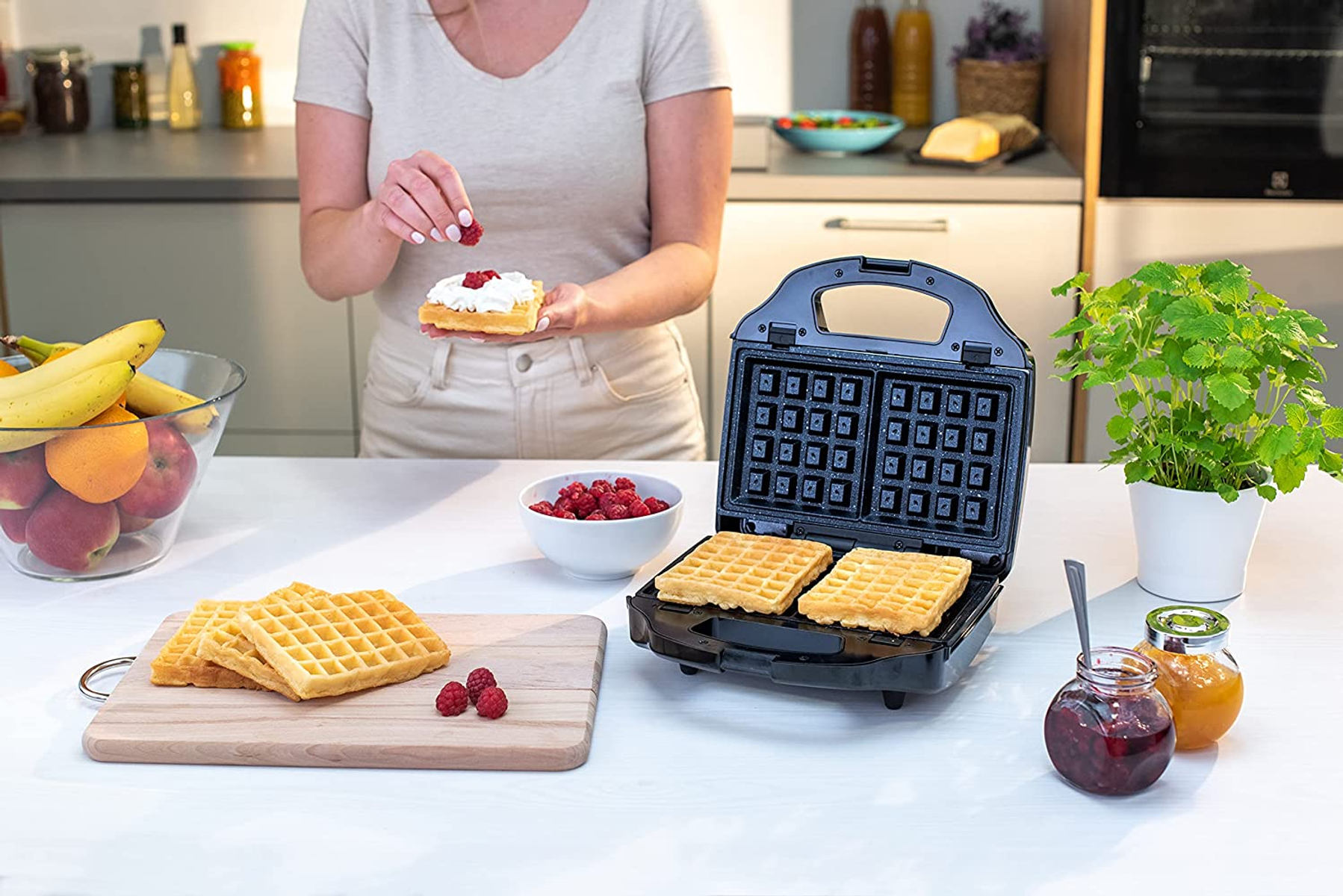 Una persona che prepara waffle con frutta e marmellata, usando una macchina per waffle.