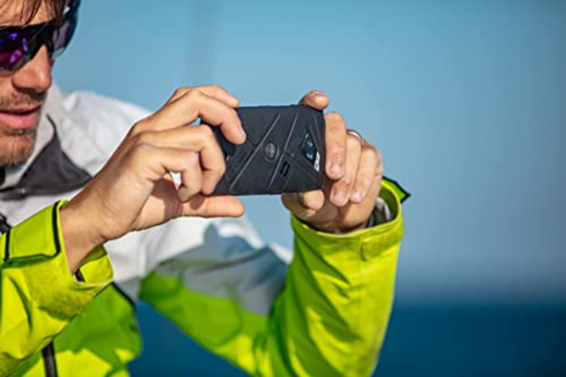 Ein Mann hält ein schwarzes Telefon mit robustem Design. Er trägt eine Sonnenbrille und eine gelbe Jacke.