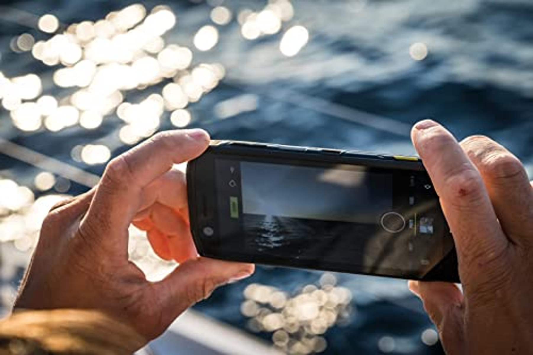 Hände halten ein schwarzes Telefon und machen ein Foto vom Meer. Sonnenlicht reflektiert auf dem Wasser.
