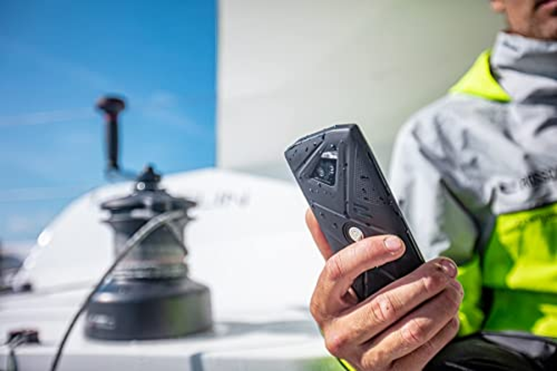 Eine Person auf einem Boot hält ein schwarzes Telefon. Der Himmel ist blau, und eine Seilwinde ist im Hintergrund.