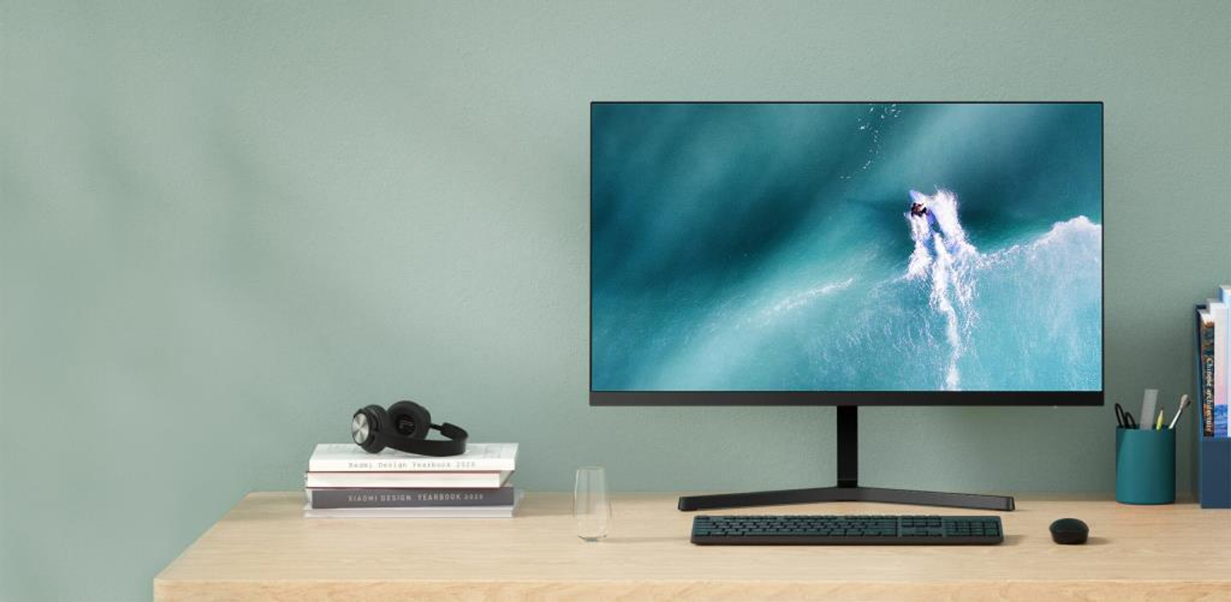 Un monitor mostra un surfista sulle onde. Una scrivania con libri, cuffie, un bicchiere e un mouse.
