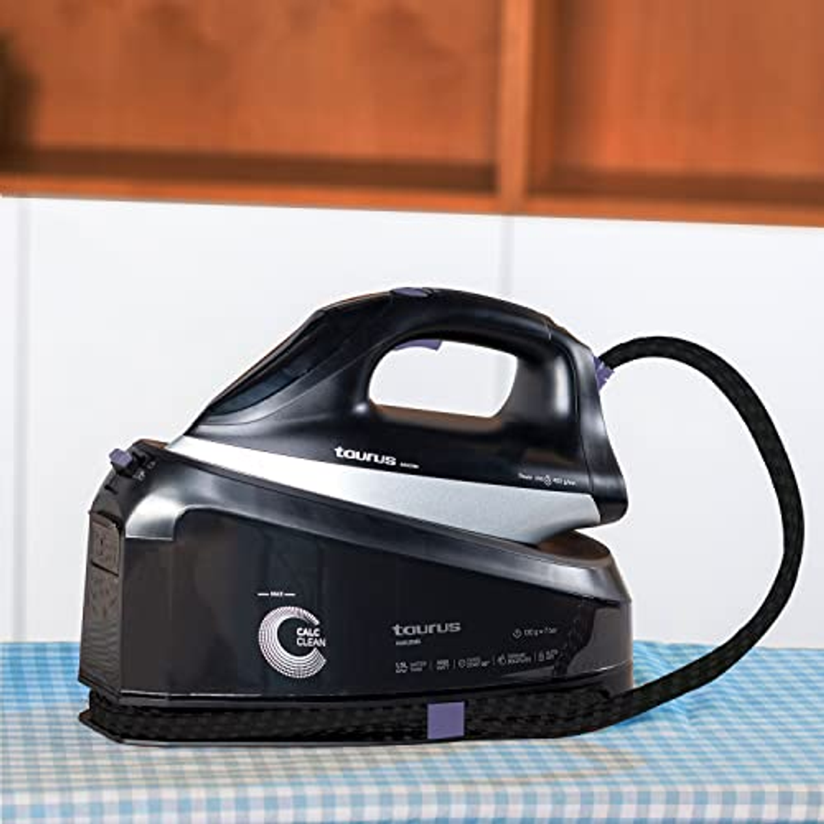 Schwarzes Taurus-Bügeleisen mit schwarzem Kabel auf blau-weiß kariertem Tisch, Wassertank.