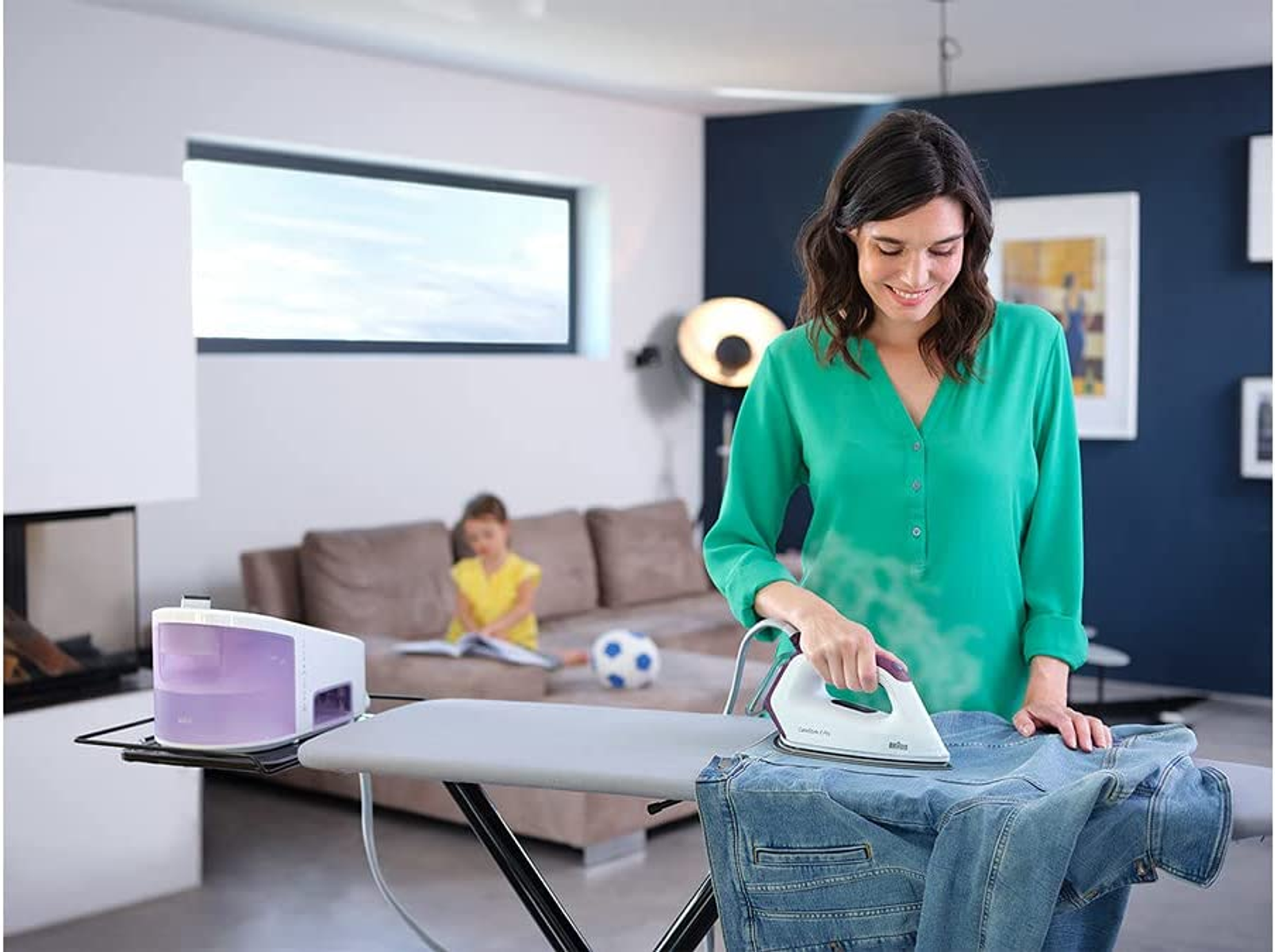 Donna che stira jeans su un'asse da stiro, con ferro a vapore e un bambino.