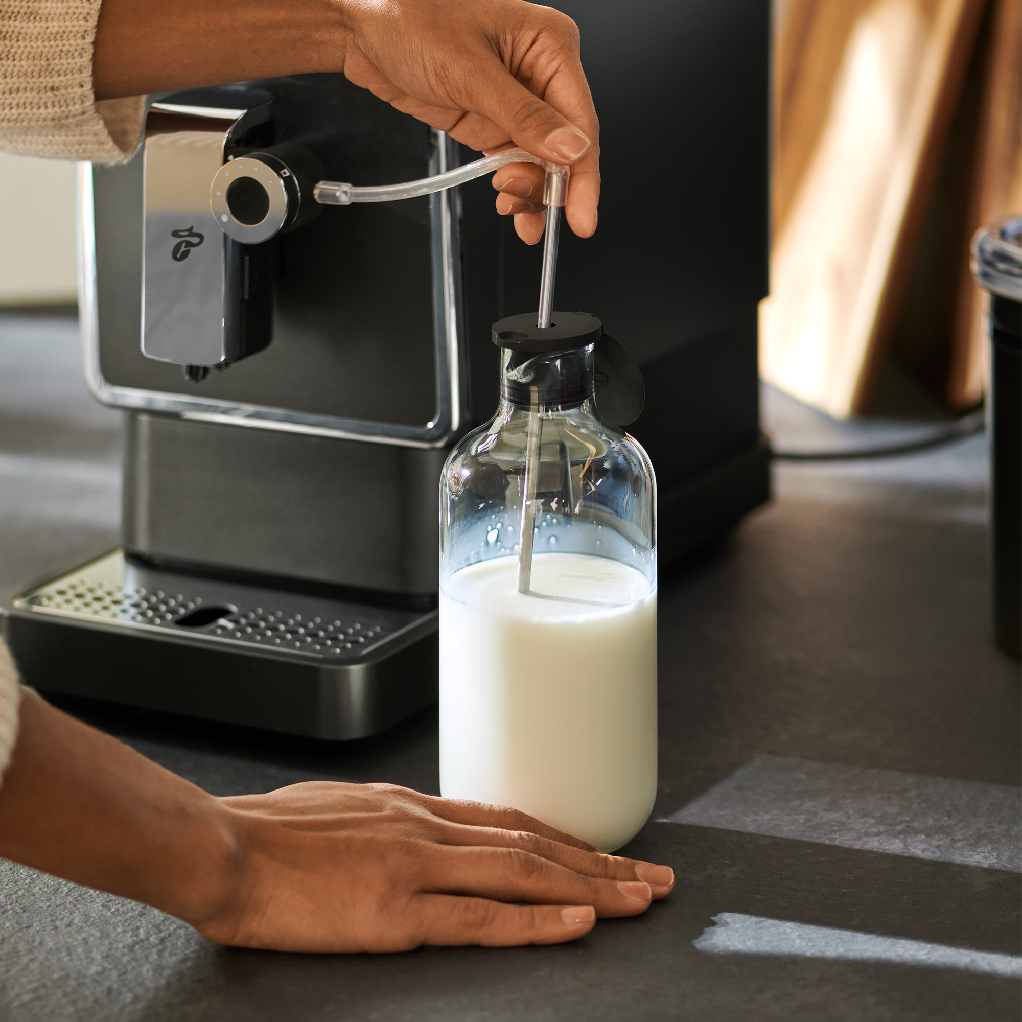 Person gießt Milch in eine Glasflasche. Espressomaschine im Hintergrund.