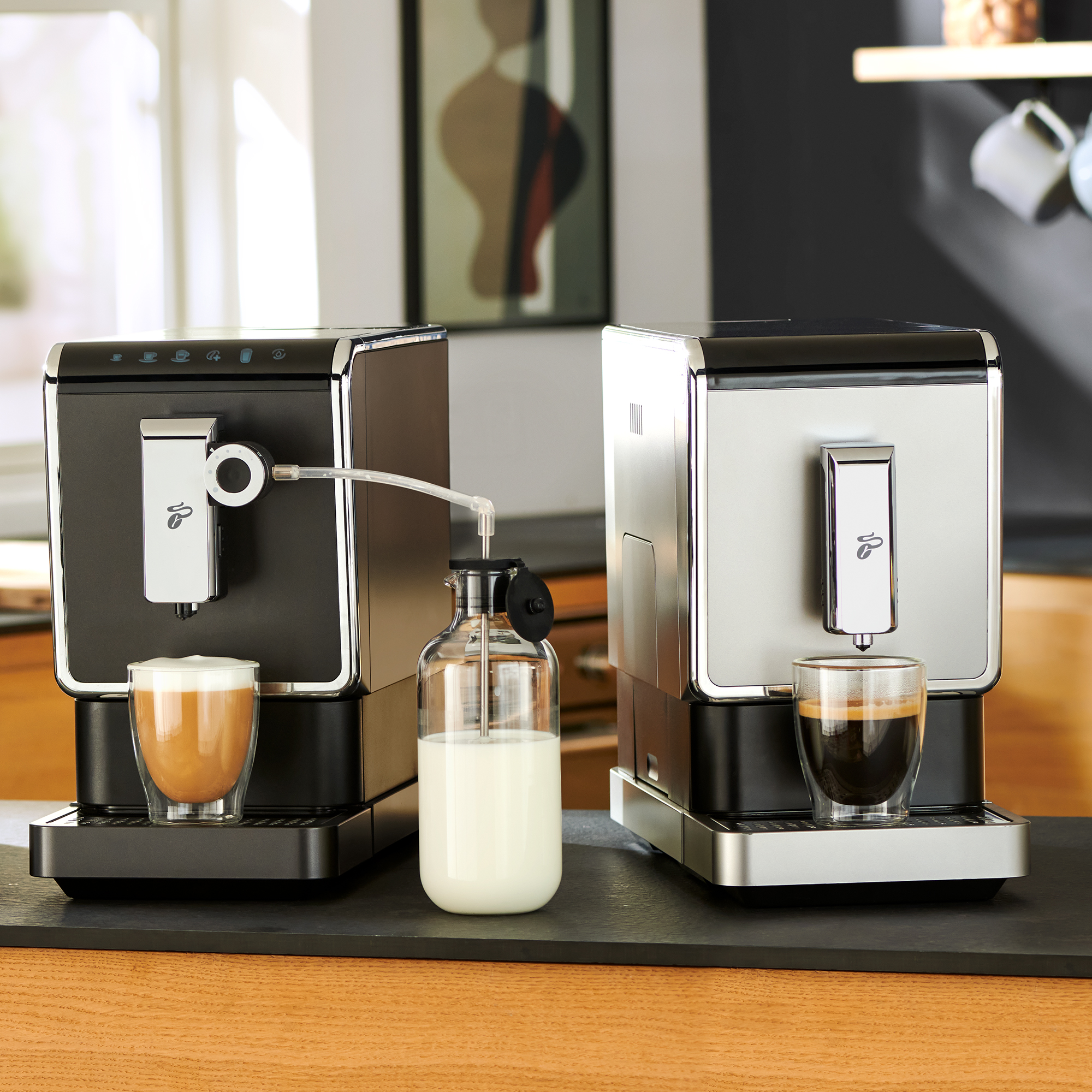 Zwei Kaffeemaschinen, eine schwarz, eine silberfarben, mit Kaffee- und Milchbechern. Zwischen ihnen steht eine Milchflasche.