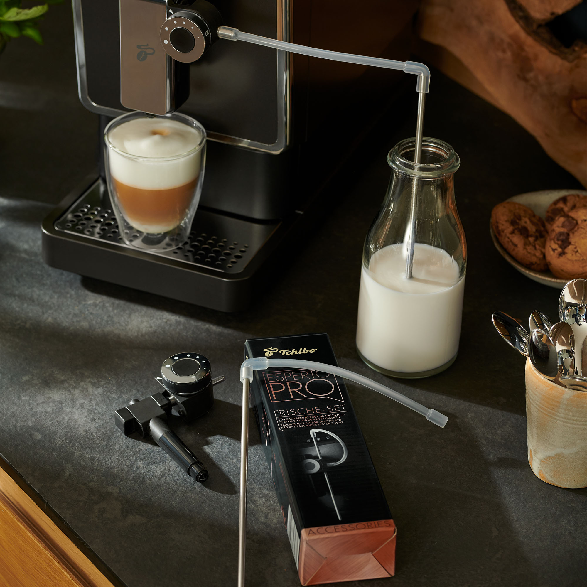 Espressomaschine mit Latte, Milchflasche und Zubehör. Kekse rechts. Schwarze Arbeitsplatte.
