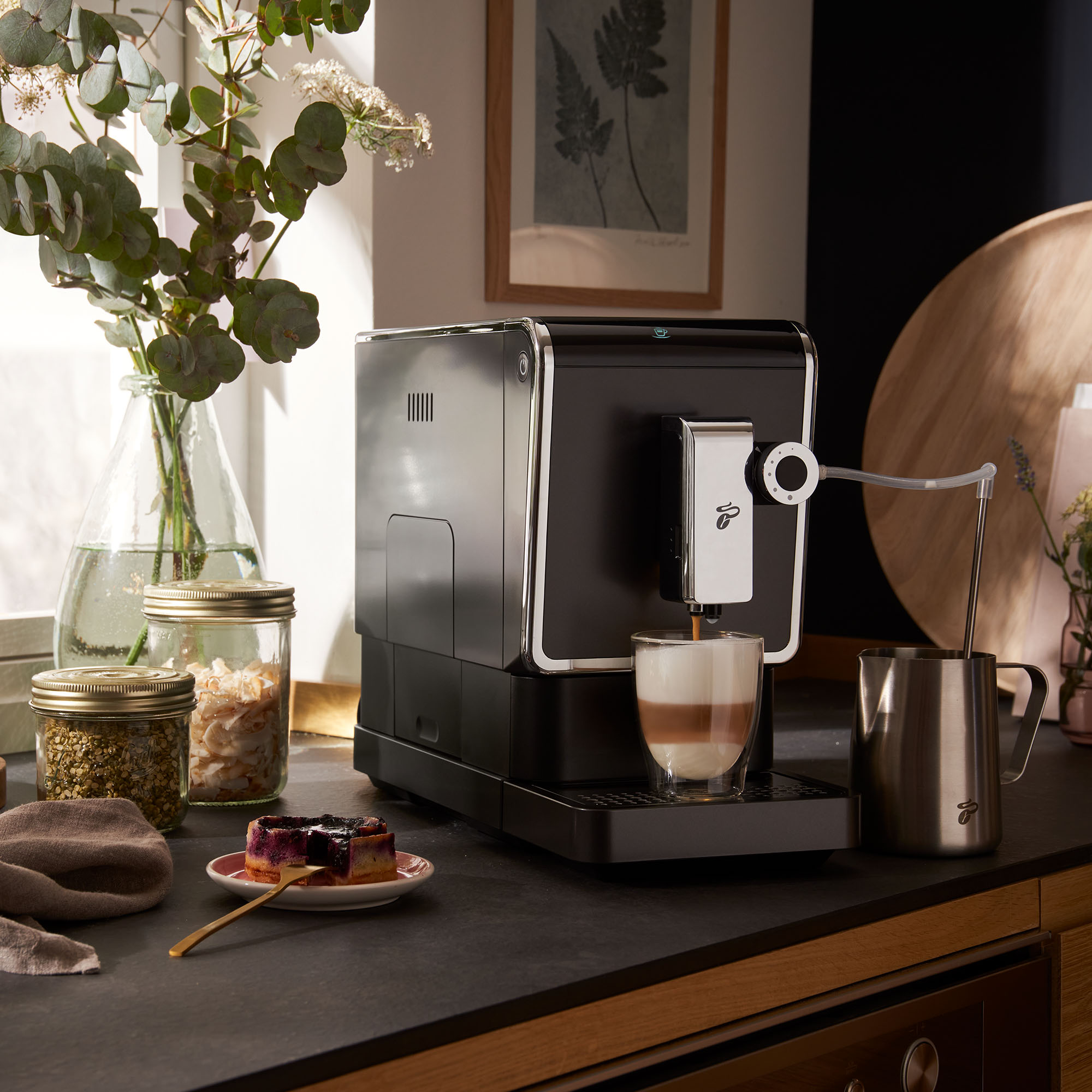 Kaffeemaschine mit Glas Latte, Stahlkrug und Kuchen auf einer Küchentheke.