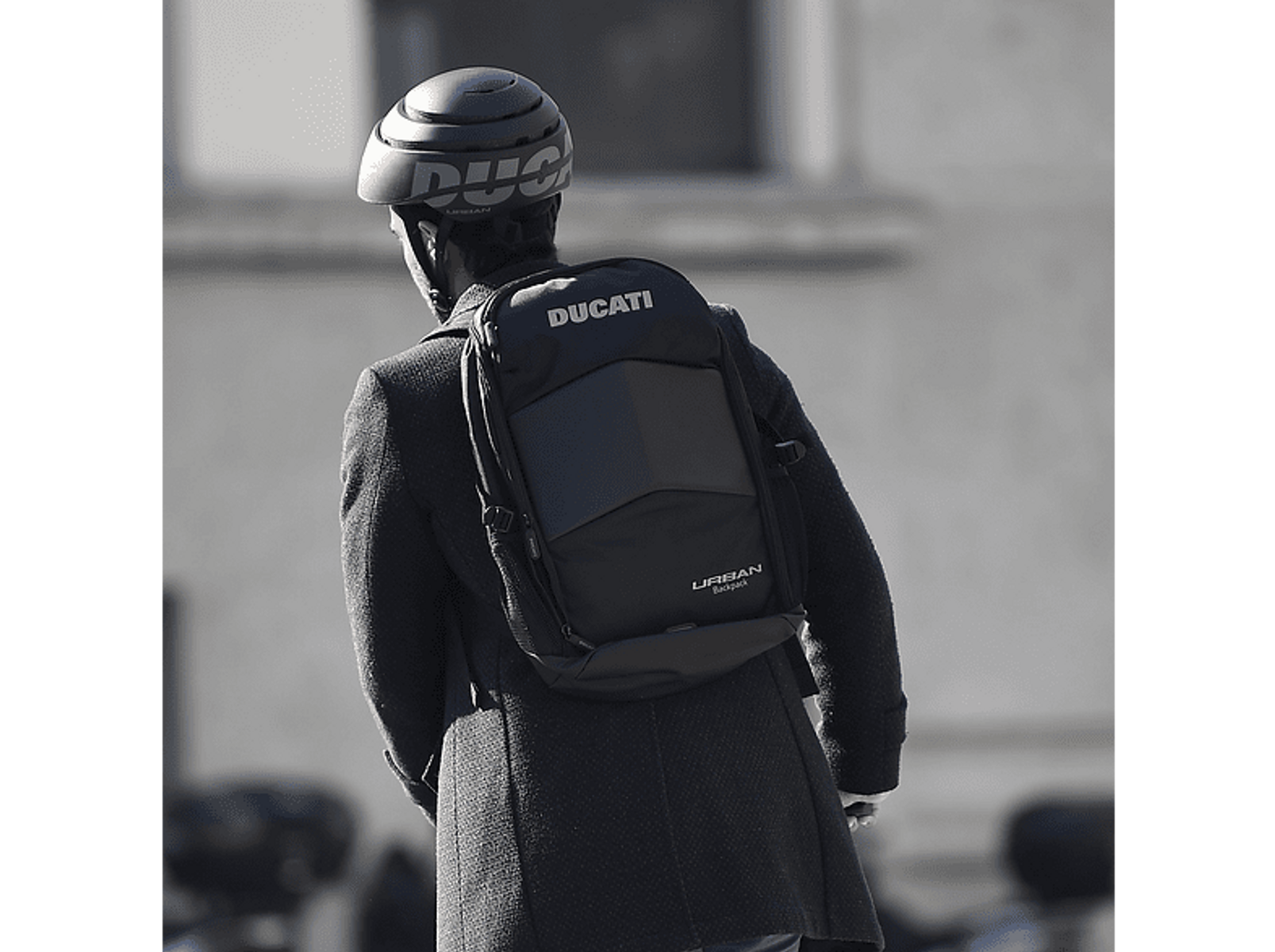 Osoba w kasku i plecaku Ducati, widziana od tyłu, idąca ulicą.