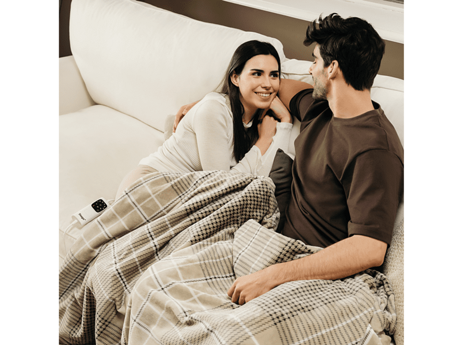 Para na kanapie, przykryta kocem. Uśmiechają się. Pokój jest dobrze oświetlony.