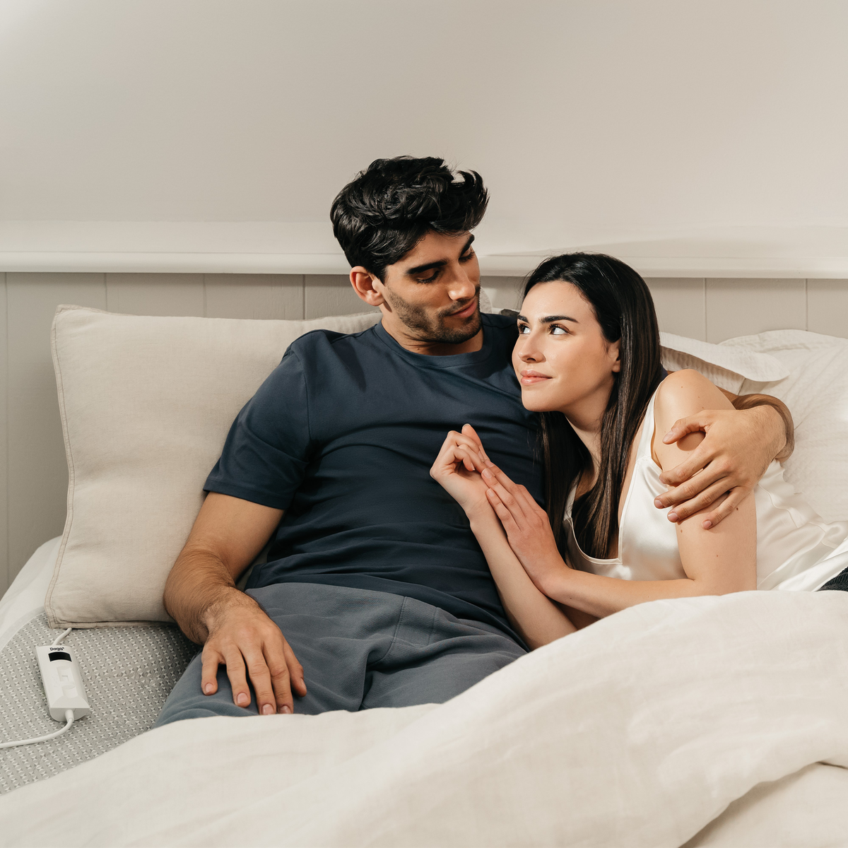 Pareja en la cama, hombre abrazando a mujer. Ambos se miran. Ropa de cama blanca, pared clara.