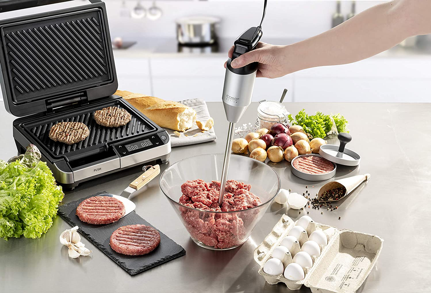 Ein Grill mit Burgern, ein Handmixer in einer Fleischschüssel und Zutaten auf einem Tisch.
