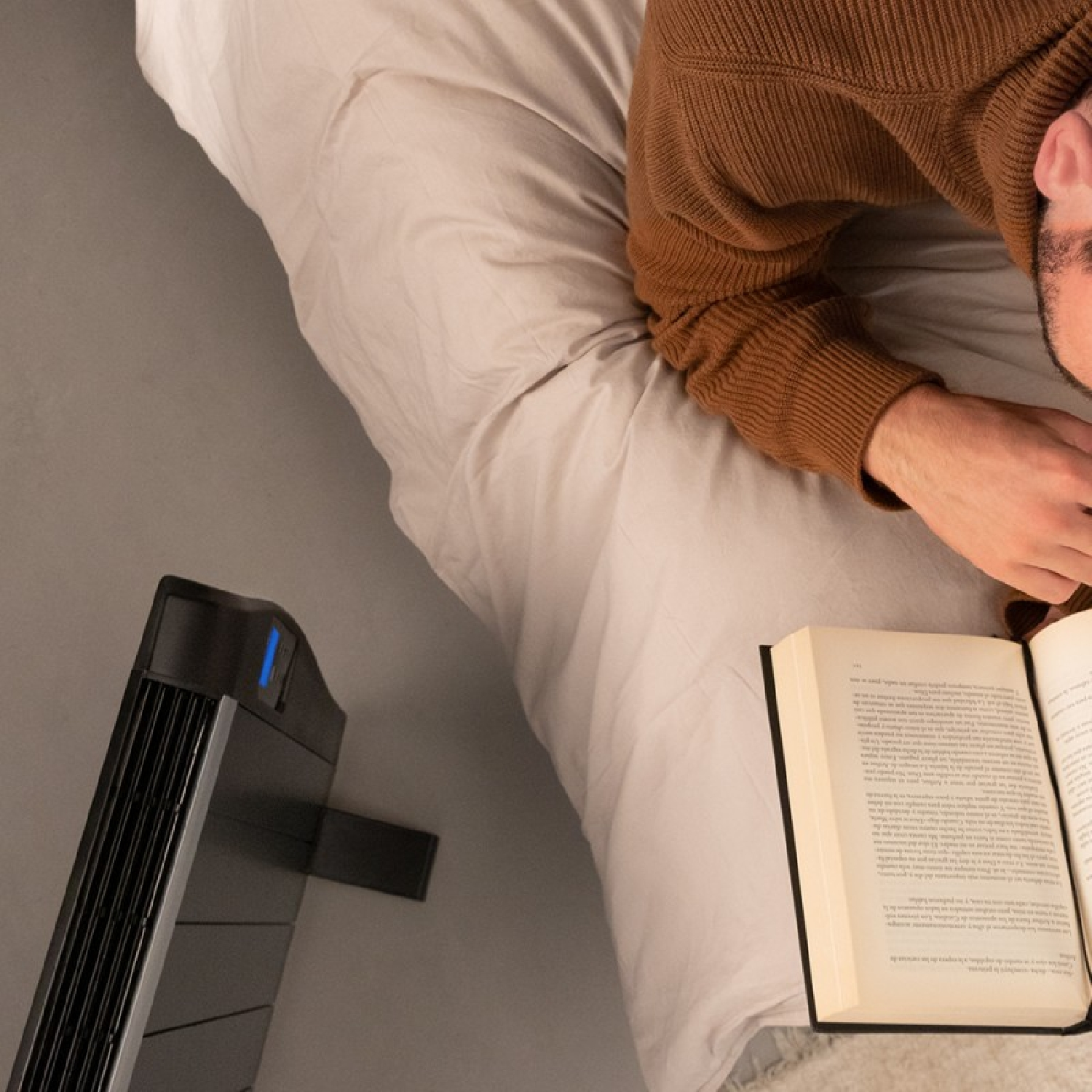 Mężczyzna czytający książkę w łóżku, obok czarnego oczyszczacza powietrza, beżowy koc.