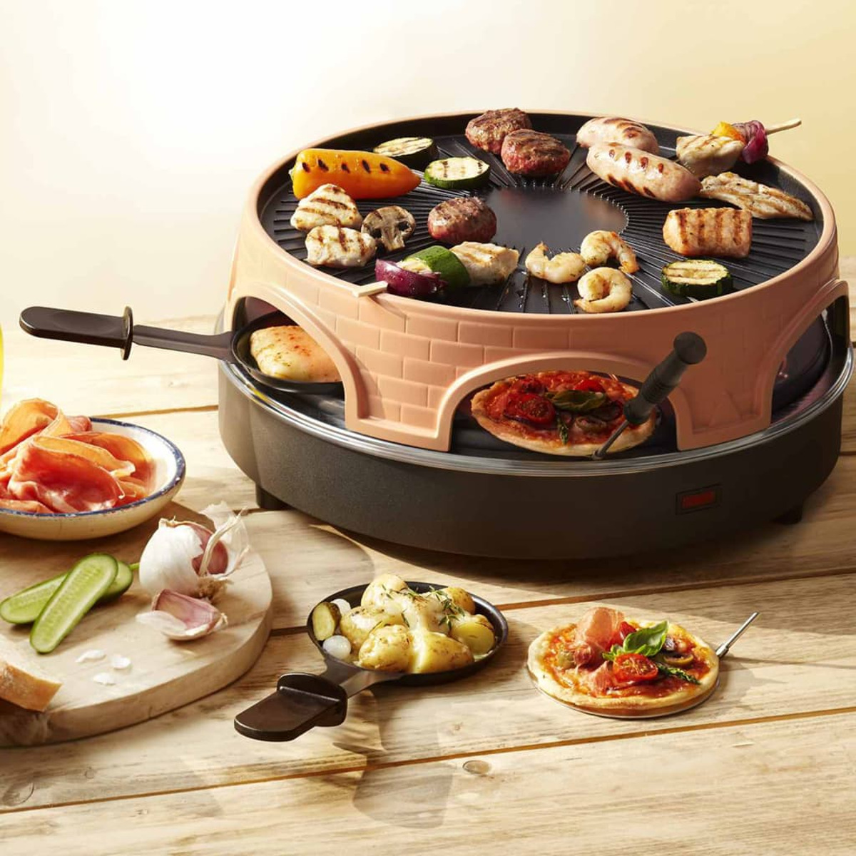 Raclette-Grill mit verschiedenen Lebensmitteln: Würstchen, Gemüse, Fleisch und Garnelen. Pizza und Kartoffeln in den unteren Schalen. Holztisch.