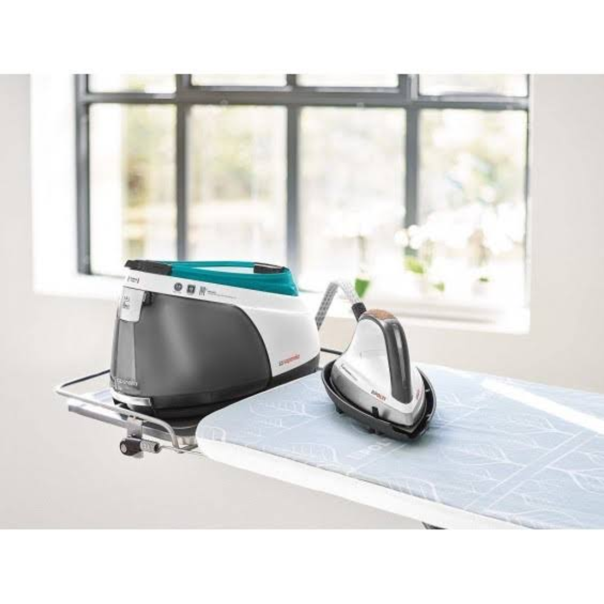 Bügelmaschine auf einem Tisch. Bügeleisen und Bügelbrett vor dem Fenster.