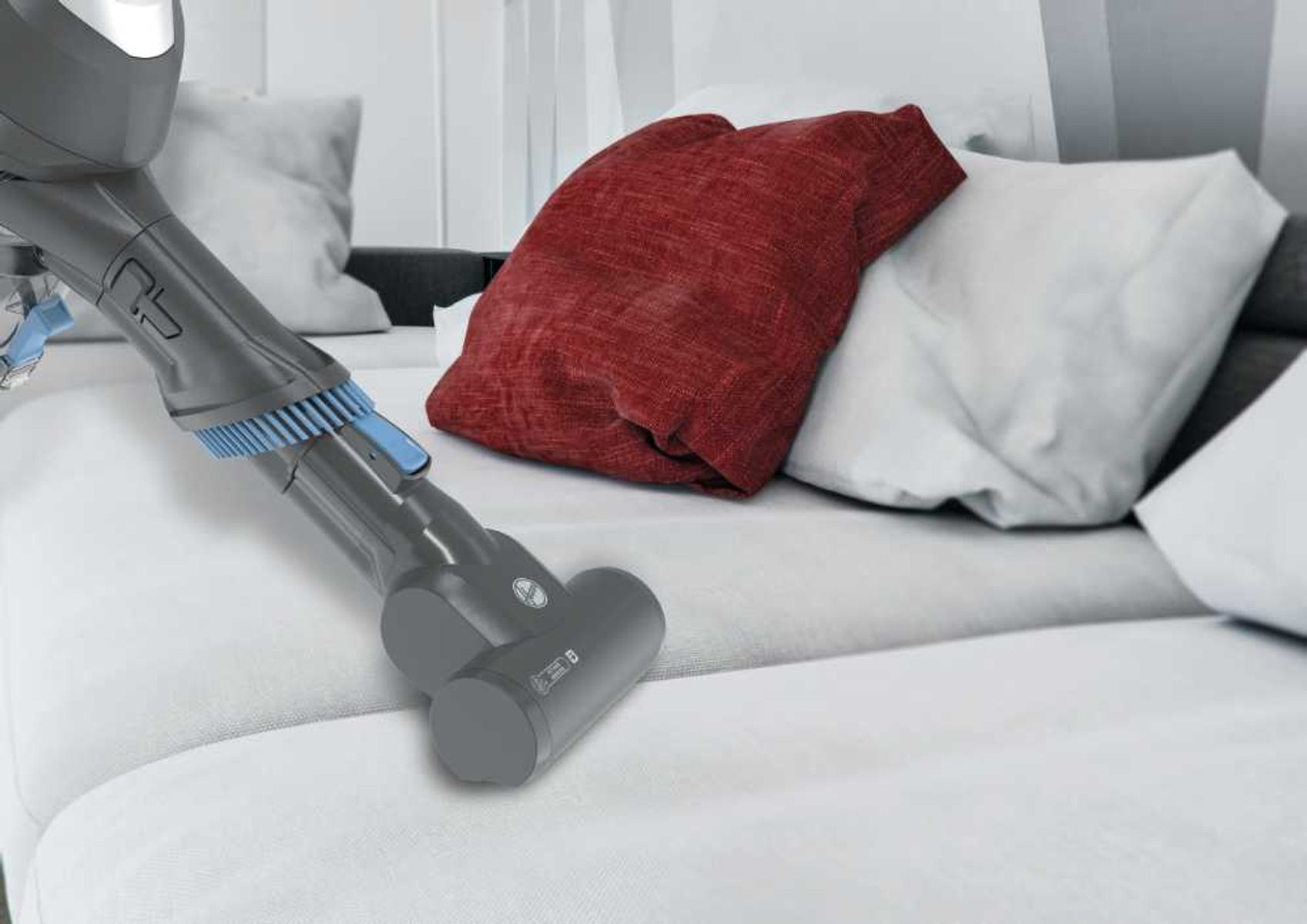 Persona aspirando un sofá blanco con aspiradora gris y azul, almohada roja visible.