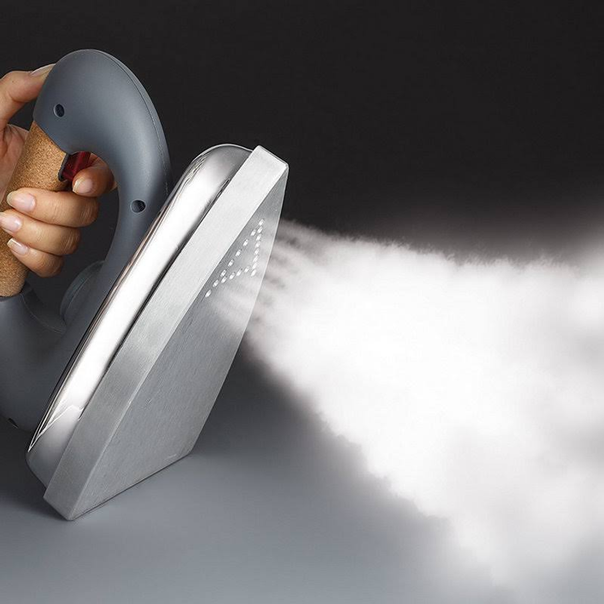 Plancha soltando vapor, sujeta con la mano, mango gris, sobre superficie gris, fondo oscuro.
