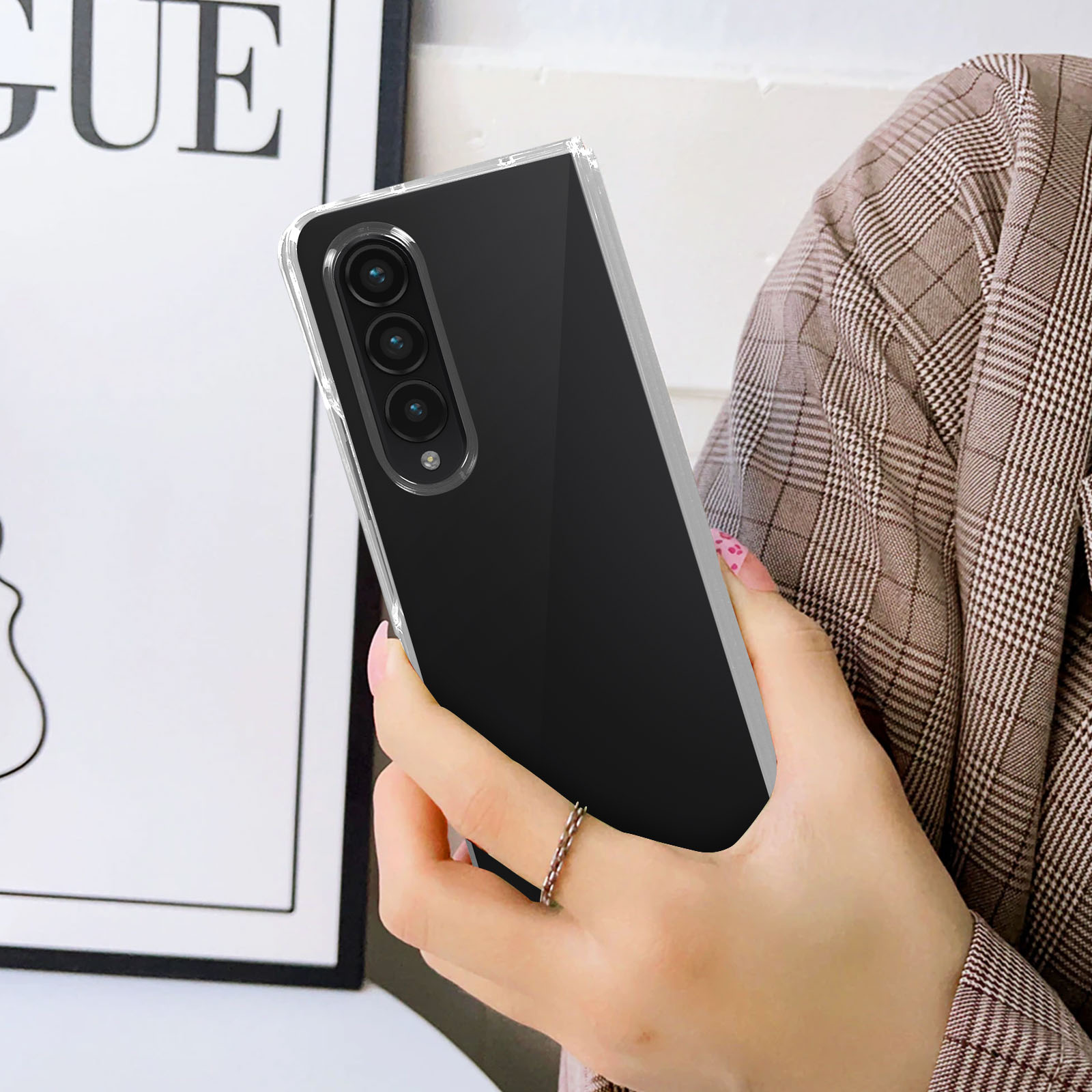Een persoon houdt een zwarte telefoon vast in een doorzichtige hoes. De telefoon heeft drie camera's. Ze draagt een blazer en een ring.