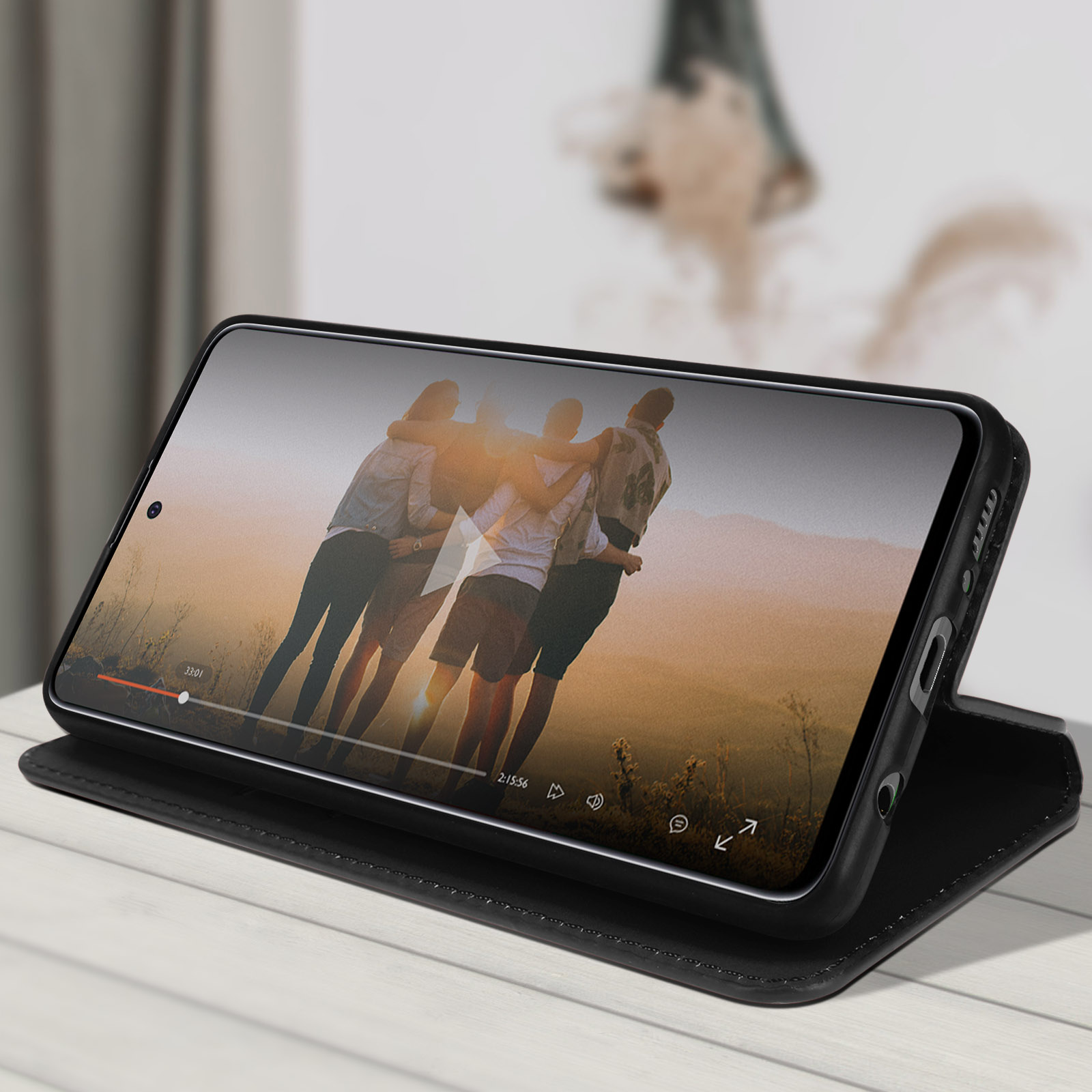 Telefon in einer Hülle zeigt Video von umarmenden Personen. Schwarze Hülle, weißer Tisch, verschwommener Hintergrund.