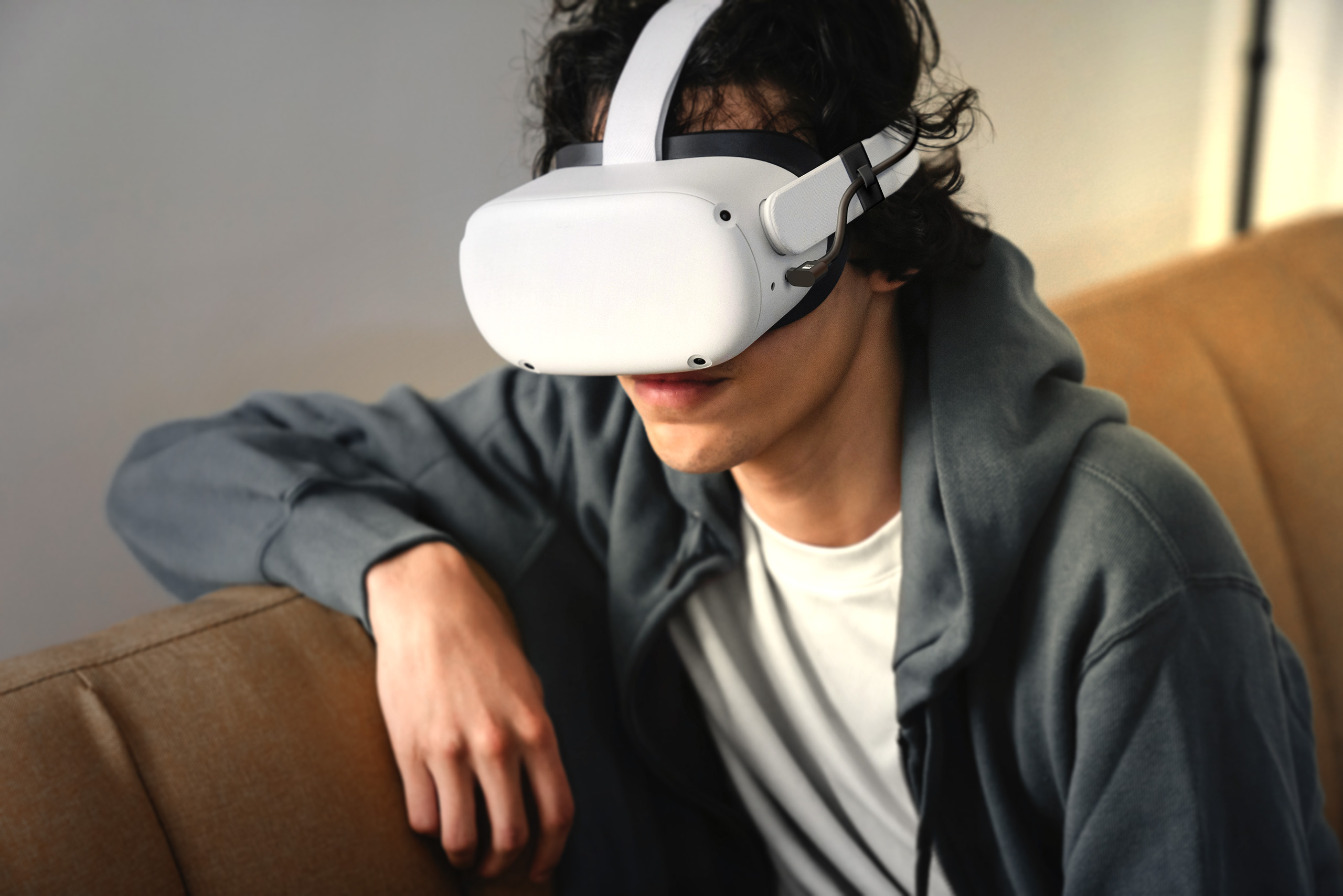 Person trägt VR-Headset, konzentriert sich auf die Erfahrung. Braunes Sofa im Hintergrund. Natürliches Licht.