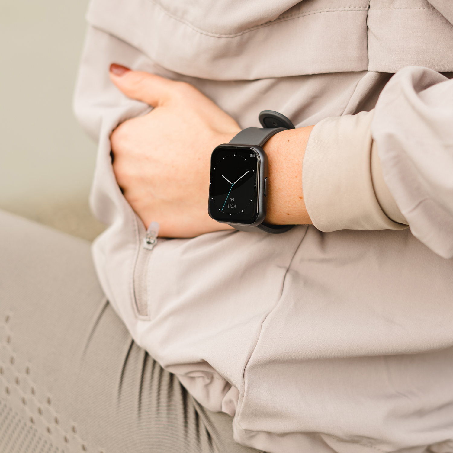 Frau trägt eine schwarze Smartwatch mit schwarzem Armband. Graue Jacke. Hintergrund ist neutral.