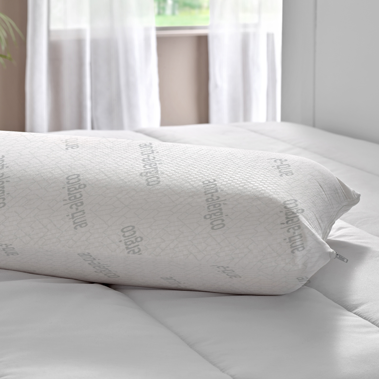 Almohada blanca y larga sobre una cama. Tiene la palabra 'anti-alergico'.