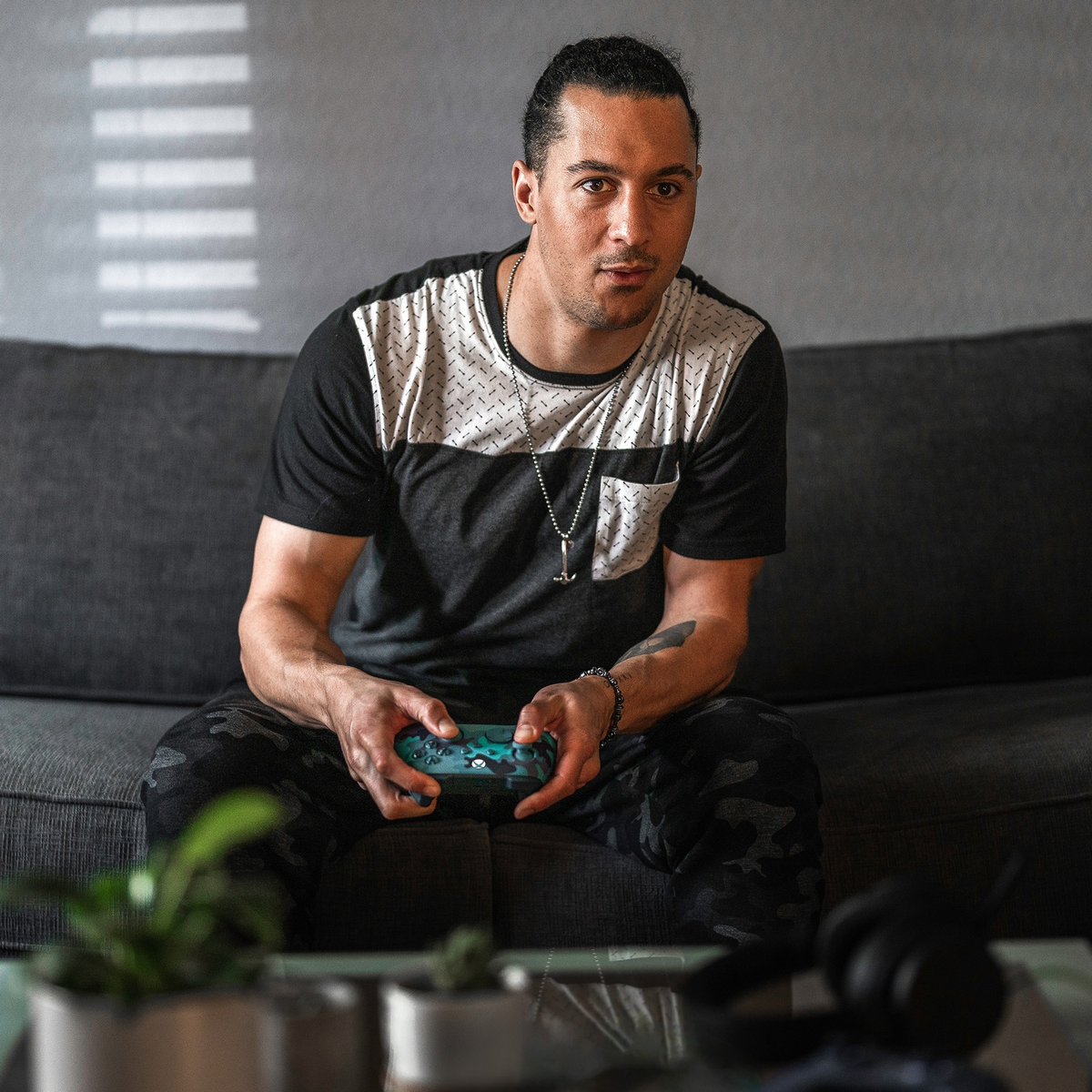 Mann sitzt auf Sofa, hält Gamecontroller. Hintergrund: Fensterjalousien und Wand.