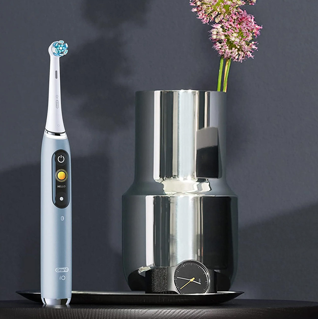 Blaue elektrische Zahnbürste neben einer silbernen Vase mit Blumen und einer Uhr auf einem schwarzen Tablett.