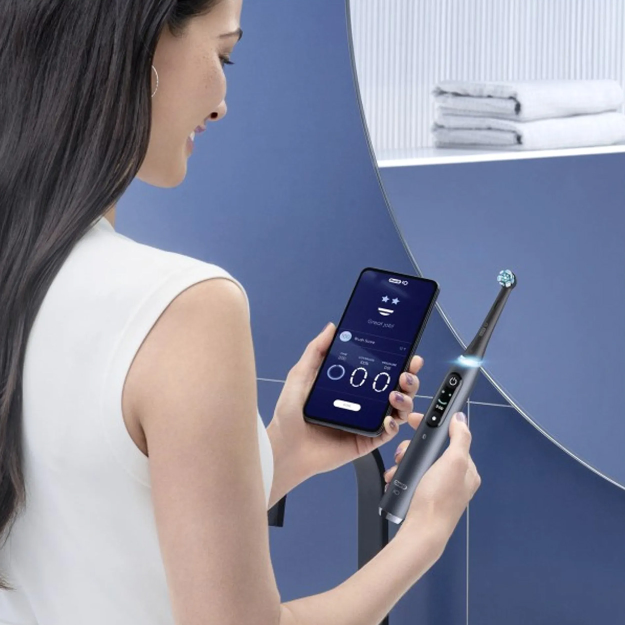 Kobieta trzymająca smartfon i elektryczną szczoteczkę do zębów przed lustrem.