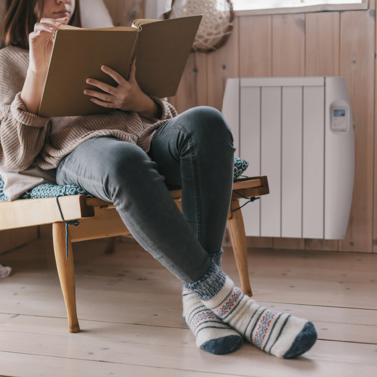 Persona leyendo un libro cerca de un calefactor, sentada en un banco de madera, habitación acogedora.