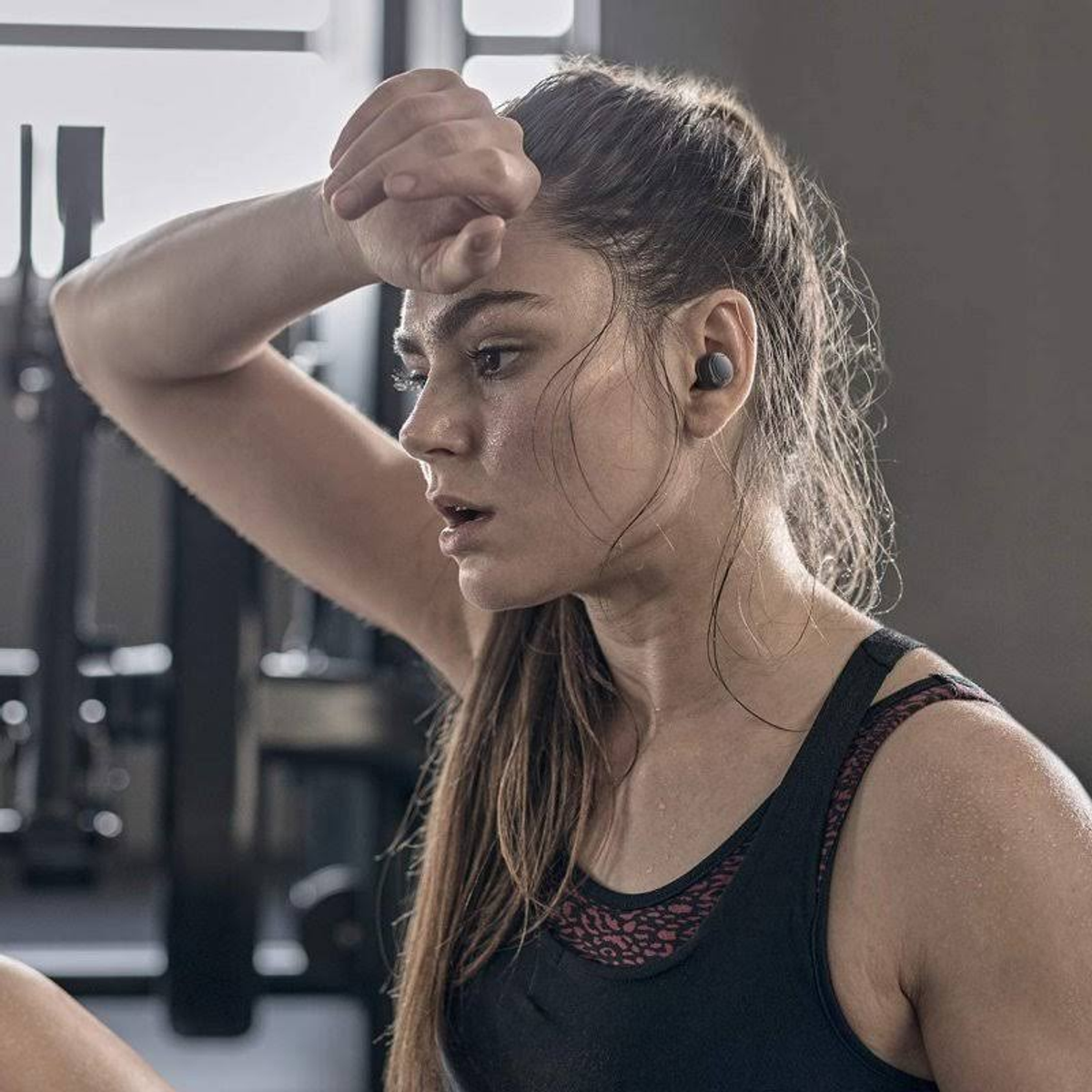 Eine Frau in Sportkleidung wischt sich den Schweiß von der Stirn, trägt Ohrhörer. Gymnastikhintergrund.