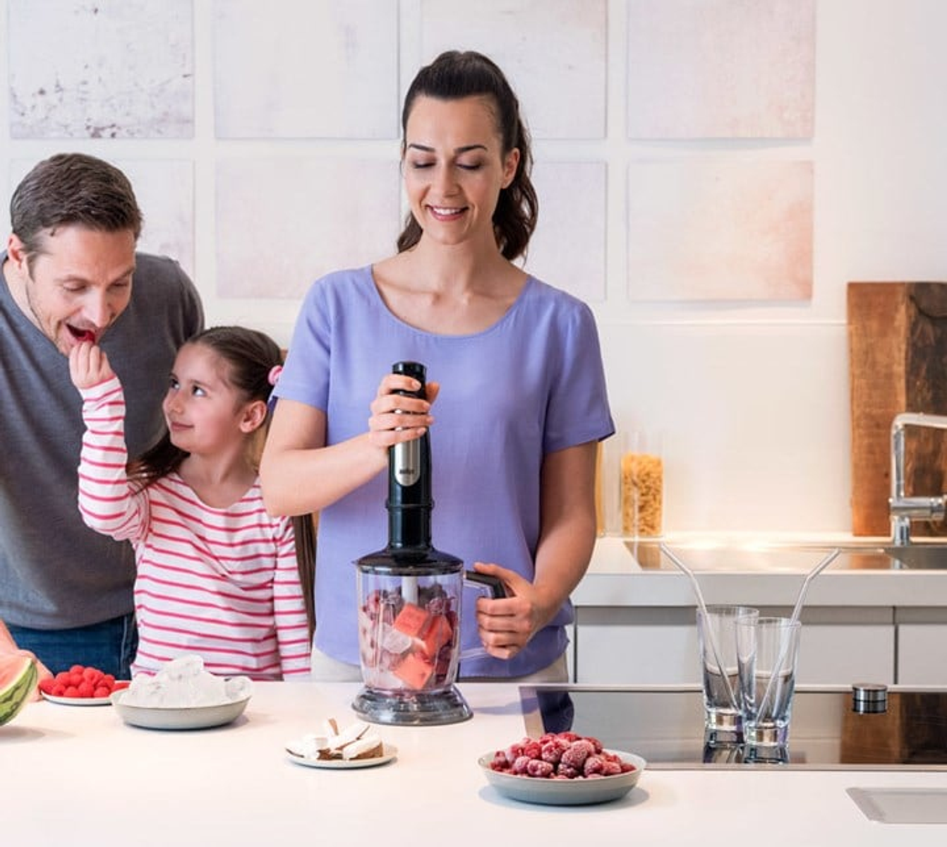 Rodzina, kobieta używająca blendera z owocami i inne owoce na stole.