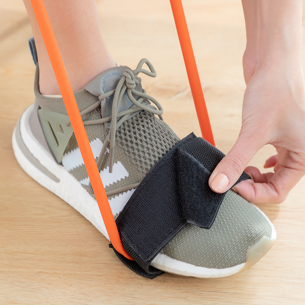 Ein Fuß in einem grünen Schuh mit schwarzem Riemen. Ein orangefarbenes Widerstandsband wird gehalten, und eine Hand sichert den Riemen.