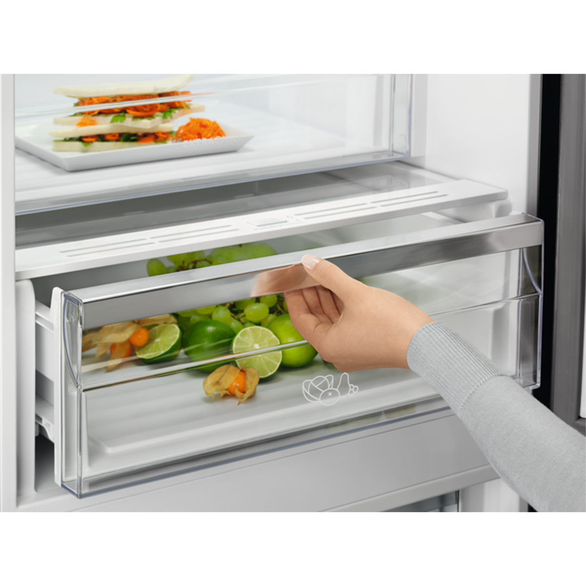 Mano abriendo un cajón de refrigerador lleno de uvas verdes, limas y otros productos.