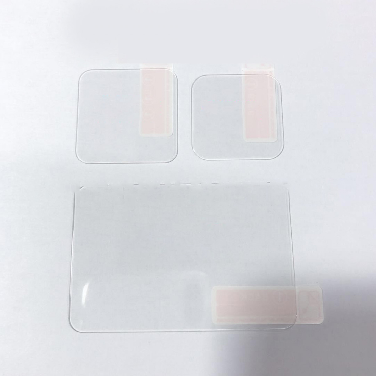 Tres protectores de pantalla con tiras adhesivas. Fondo blanco. Hojas de plástico transparente.