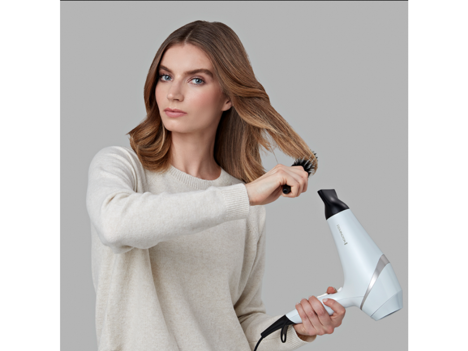 Kobieta szczotkuje włosy okrągłą szczotką, trzymając białą suszarkę do włosów.