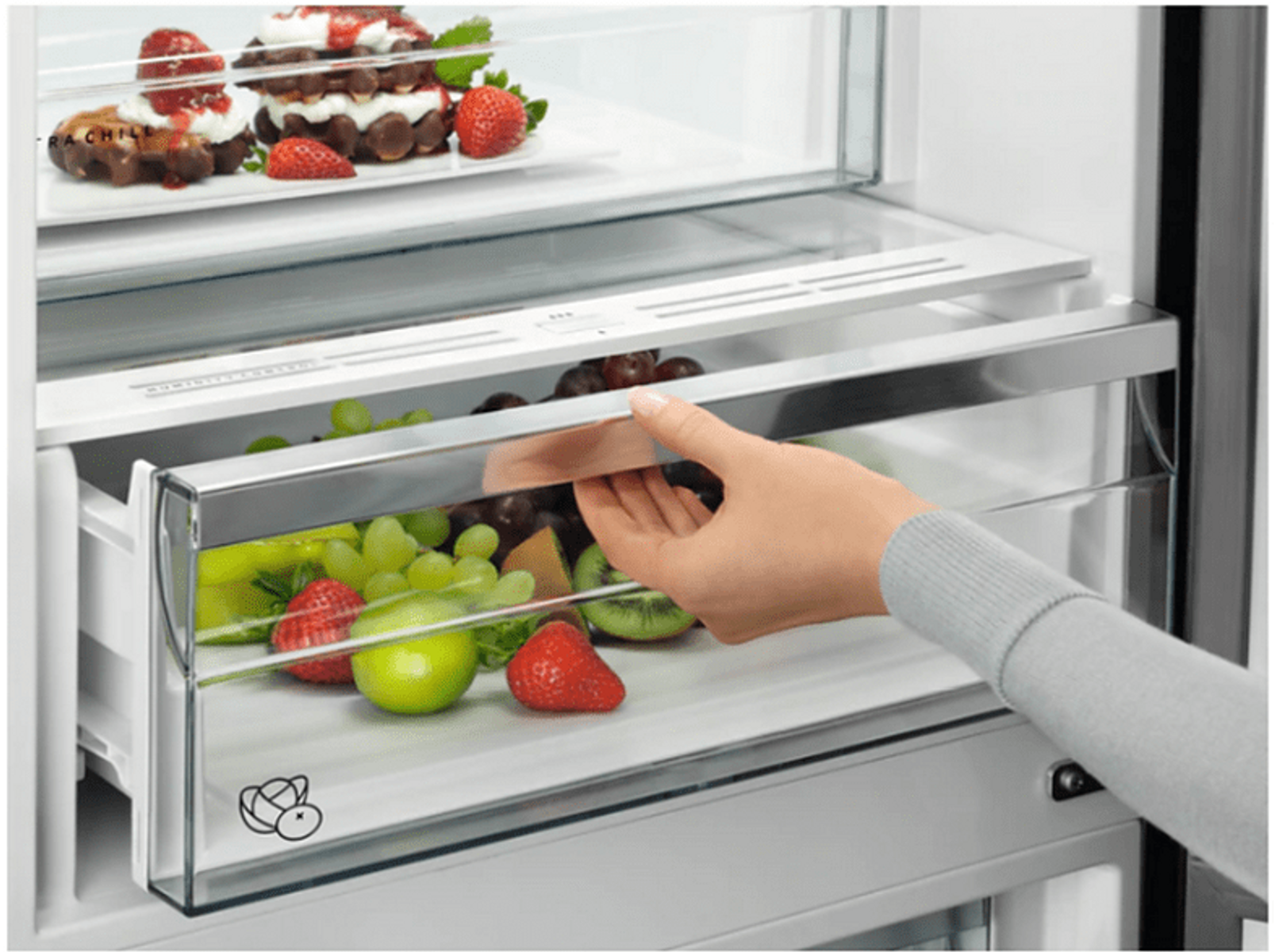 Ręka otwiera szufladę lodówki z owocami i deserem na górnej półce.