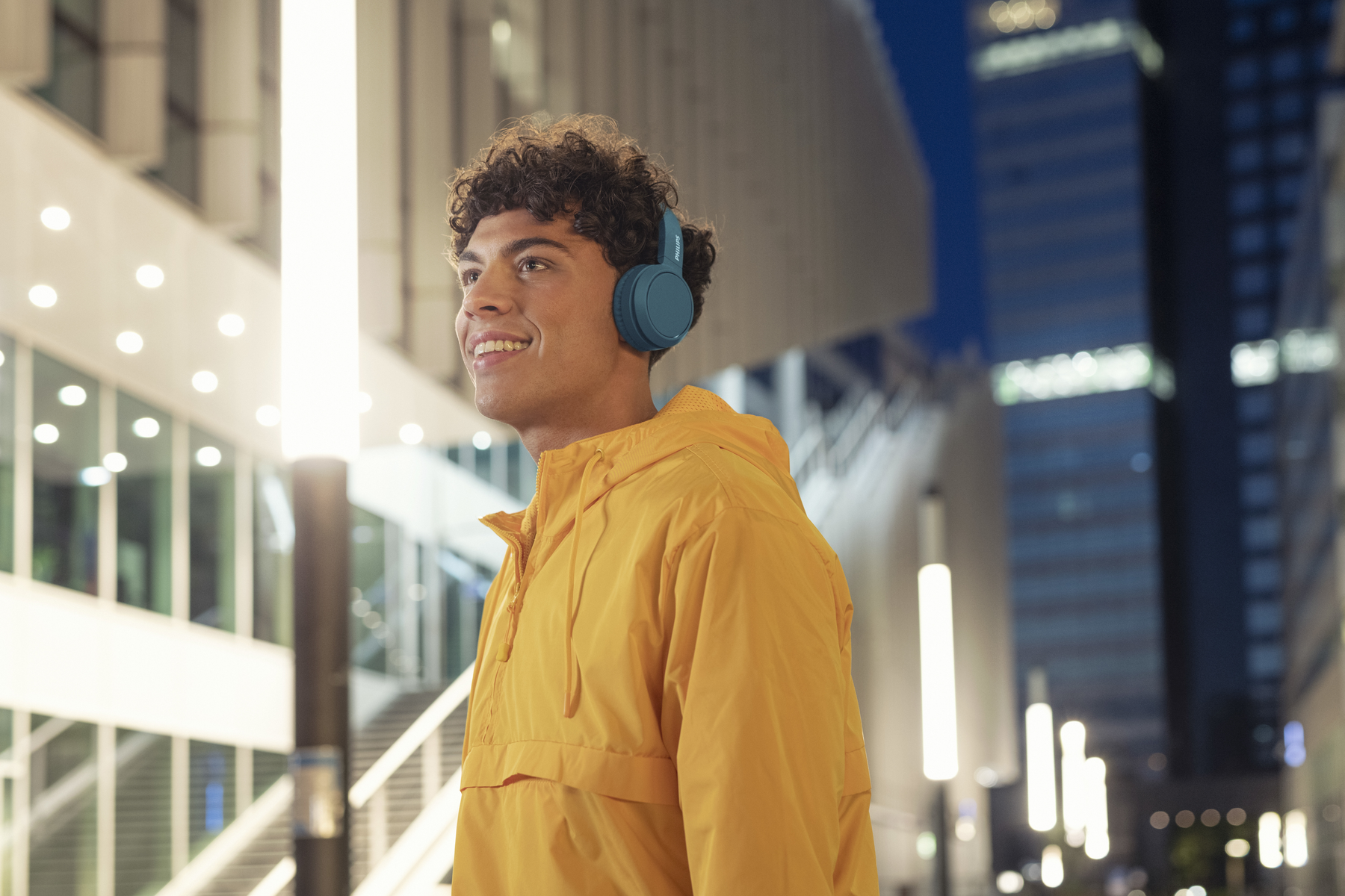 Joven con cabello rizado con auriculares y chaqueta amarilla sonríe al aire libre.