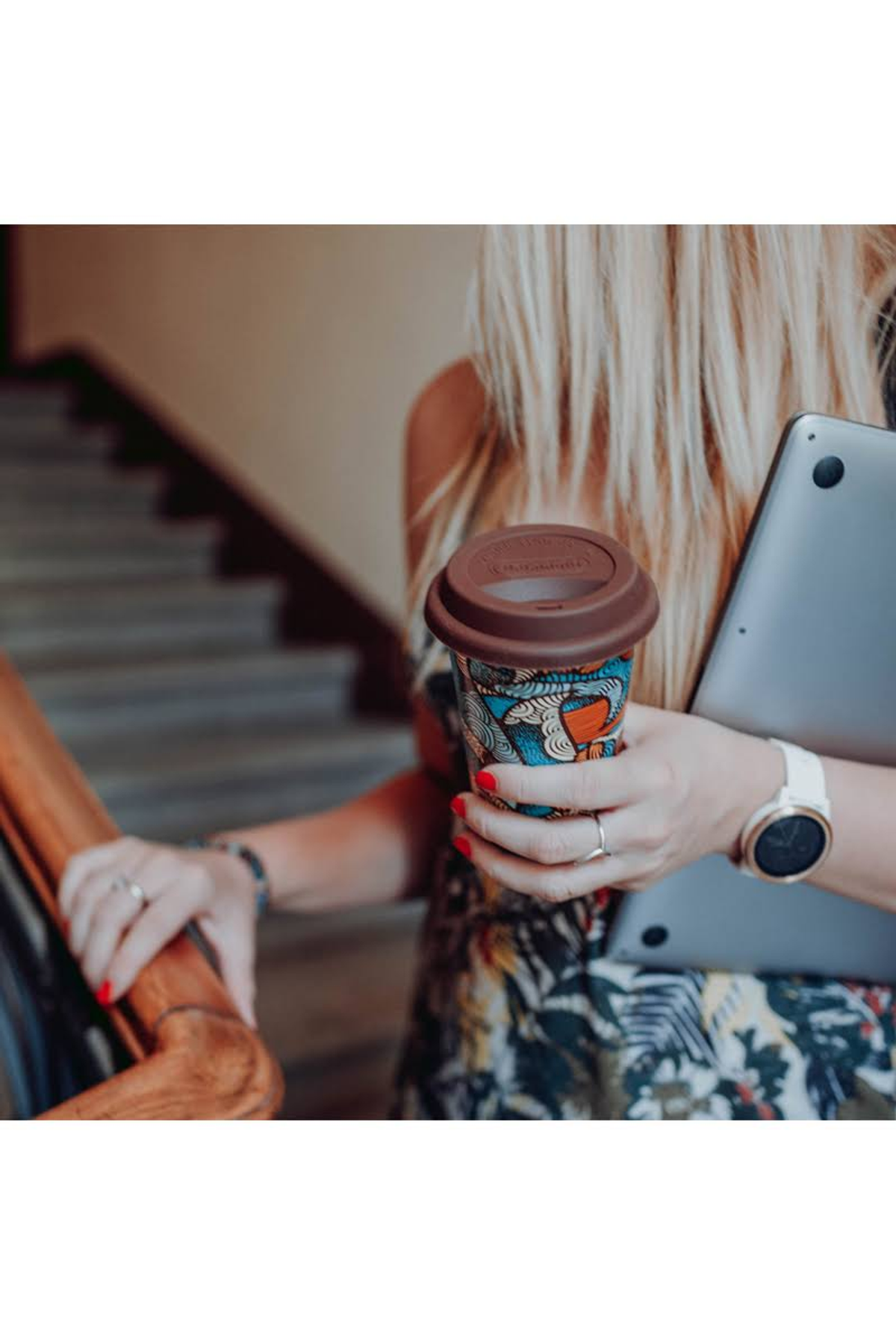 Mujer rubia sosteniendo una taza de café y un portátil, subiendo escaleras, usando reloj y anillos.