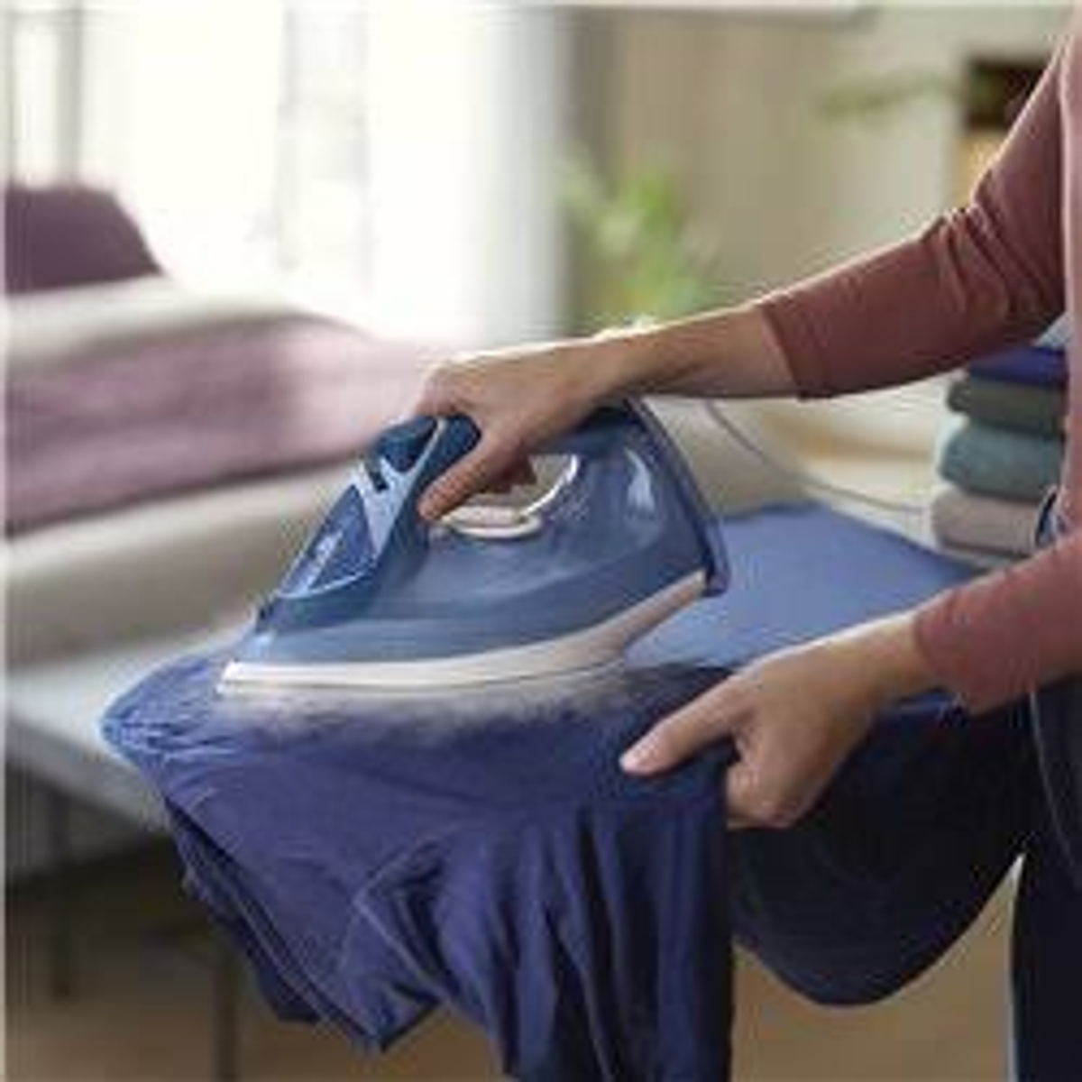Personne repassant une chemise bleue sur une planche. Vapeur visible. Arrière-plan flou.