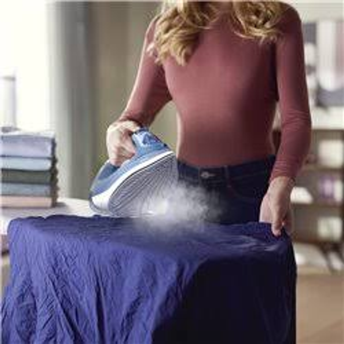 Femme repassant une chemise bleue. De la vapeur sort du fer à repasser. Une pile de serviettes est à proximité.