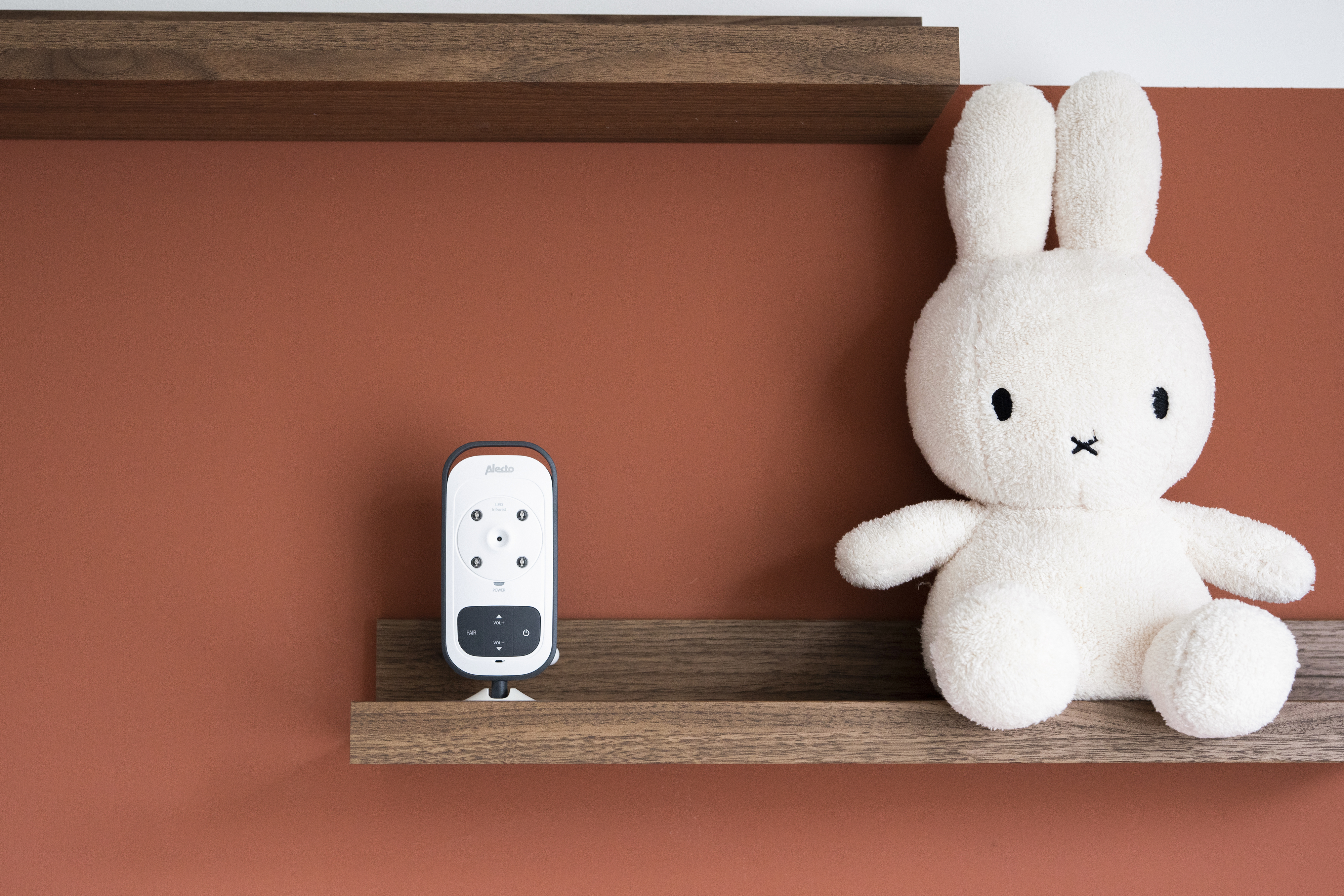 Niania elektroniczna i pluszowy królik na półce na tle czerwonobrązowej ściany. Tło jest rozmyte.